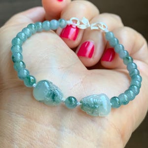 Puede incluir: Una pulsera de cuentas con pequeñas esferas translúcidas de color verde jade claro y dos cuentas más grandes talladas con forma de animal en el mismo tono verde. La pulsera se asegura con un cierre de metal plateado y está ensartada en un cordón fino.
