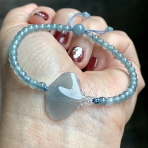 Może przedstawiać: Jasnoniebieska bransoletka z koralików z zawieszką w kształcie serca. Bransoletka posiada małe, okrągłe koraliki i niebieskie sznurkowe zapięcie. Zawieszka jest przezroczysta i blado niebieska. Bransoletka jest trzymana w dłoni.