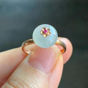 May include: A delicate ring with a round, light green jade stone set in a rose gold band. A small gold flower with a red center sits atop the stone.