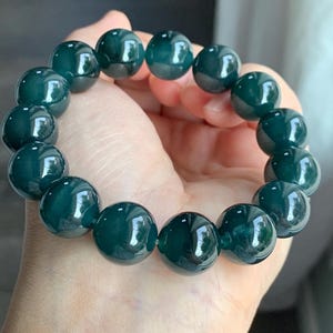 May include: A dark teal beaded bracelet. The bracelet is made of large, round, polished beads. The beads have a glossy finish and reflect light. The bracelet is worn on a wrist.
