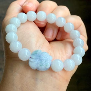 Puede incluir: Una pulsera de jade blanco con cuentas grandes y redondas. La pulsera presenta una pieza central floral tallada, de color azul claro y verde. Las cuentas están ensartadas en un cordón elástico morado. La pulsera se sostiene en una mano.