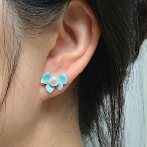 May include: A close-up of a flower-shaped earring. The earring features a light blue enamel flower design with a small, round, white pearl in the center. The earring has a gold-colored metal border and post. The earring is worn on an ear.