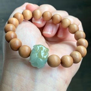 May include: A beaded bracelet featuring light brown wooden beads and a central, carved jade accent. The jade is a pale green color and is carved into a floral design. The bracelet is held in a hand.
