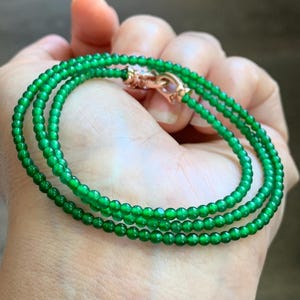 May include: A multi-strand necklace or bracelet composed of small, translucent, vibrant green spherical beads. The jewelry features a metallic rose gold-toned clasp mechanism that connects the multiple strands of beads into a single circular loop.