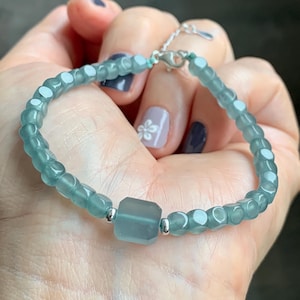 May include: A light blue beaded bracelet with a square center bead and silver accents. The bracelet is made of small, faceted beads and is held in a hand.
