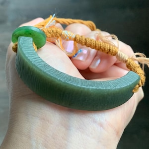 May include: A jade bracelet featuring a curved, dark green stone and a round green bead. The bracelet is connected by a woven, mustard-coloured cord.