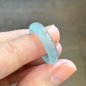 May include: A faceted, light blue ring. The ring is held between fingers, showcasing its geometric design and translucent quality. The ring's color is a soft, cool tone, suggesting a gemstone or glass material.