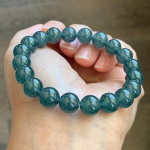 May include: A stretch bracelet composed of smooth, round, translucent dark teal beads. The beads exhibit a glossy finish and a consistent deep blue-green color throughout, arranged in a circular pattern to form a continuous loop for the wrist.