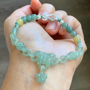 May include: A jade bracelet featuring a floral design and a dangling flower charm. The bracelet has green and yellow beads, and a silver clasp. The bracelet is held in a hand.