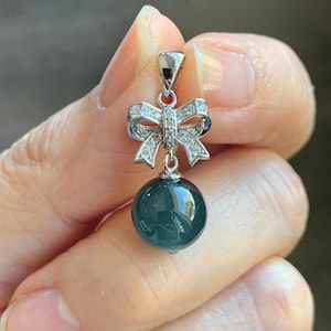 May include: A silver pendant featuring a bow design adorned with small, clear gemstones. Below the bow hangs a dark teal, spherical bead. The pendant is held in a hand against a blurred background.