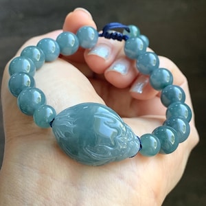 May include: A light blue beaded bracelet with a central carved oval stone. The bracelet features round beads and a detailed carved design on the oval centerpiece. The bracelet is held in a hand.