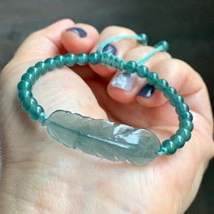 Puede incluir: Una pulsera de cuentas turquesas con un colgante de pluma tallada. La pluma es translúcida, de color verde pálido, y las cuentas son pequeñas y redondas. La pulsera es ajustable con un cierre de nudo corredizo.