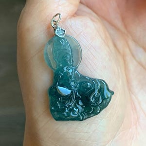 May include: A teal-colored pendant featuring a carved figure with a halo. The pendant is suspended from a silver-colored bail. The figure is detailed with flowing robes and a serene expression. The pendant is held in a hand.