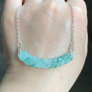 May include: A silver chain necklace with a carved jade pendant in the shape of a leaf. The pendant is a light green colour with darker green veins.