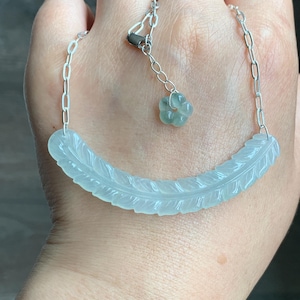 May include: A delicate necklace featuring a carved, pale green feather pendant. The feather design is detailed with etched lines, and the necklace has a silver chain with a small flower charm. The necklace is displayed on a hand.