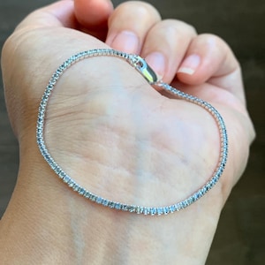 May include: A delicate silver bracelet with small, clear gemstones. The bracelet is clasped and rests gently on a person's skin, showcasing its elegant design. The gemstones sparkle, reflecting light.