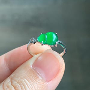 May include: A silver ring with a bright green jadeite gourd-shaped stone. A small, clear gemstone is set into the ring's band. The ring is held up against a blurred background.