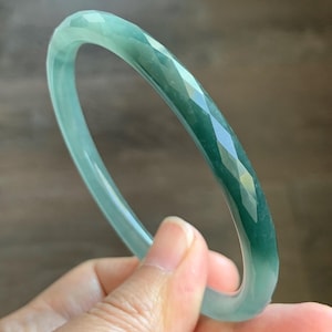 May include: A translucent green jade bangle bracelet featuring a faceted exterior surface. The circular jewelry piece displays varying shades of deep and light green with a smooth, polished finish throughout the entire circumference of the band.