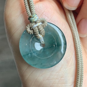 May include: A teal jade pendant with a circular design. The pendant hangs from a beige cord with a decorative knot. The jade has a polished surface, and the cord is a braided style. The pendant is a beautiful piece of jewellery.