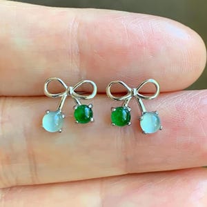 May include: A pair of silver bow earrings, each with a small, round, light blue stone and a square, vibrant green stone. The earrings are displayed against a blurred background.
