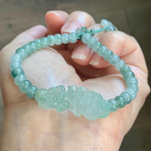 May include: A light green jade bead bracelet featuring a central carved floral motif. The bracelet is constructed with smooth, translucent, round beads and includes an adjustable light green cord closure with small matching bead accents.