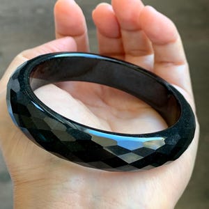 May include: A solid black bangle bracelet featuring a faceted, geometric surface texture. The circular accessory has a polished, glossy finish that reflects light across its multiple angled planes, creating a distinct pattern of dark, sharp-edged shapes.