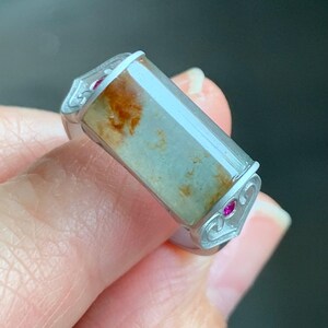 May include: A silver ring with a rectangular green and brown stone set in the center. The ring has two small red stones on either side of the stone.