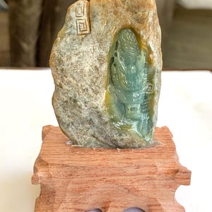 May include: A carved green and tan stone sculpture featuring a dragon relief, mounted on a rectangular wooden base. The stone has a rough, textured exterior with a polished, smooth dragon carving in the center and a small square symbol at the top.