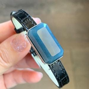 May include: A black leather bracelet with a dark blue gemstone set in a silver bezel.