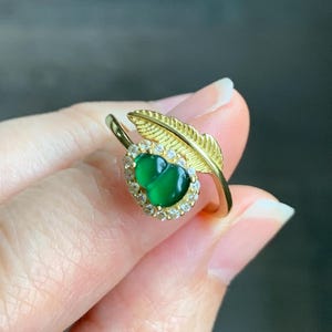 May include: A gold ring with a green jade stone set in a halo of clear cubic zirconia stones. A gold leaf design wraps around the stone.