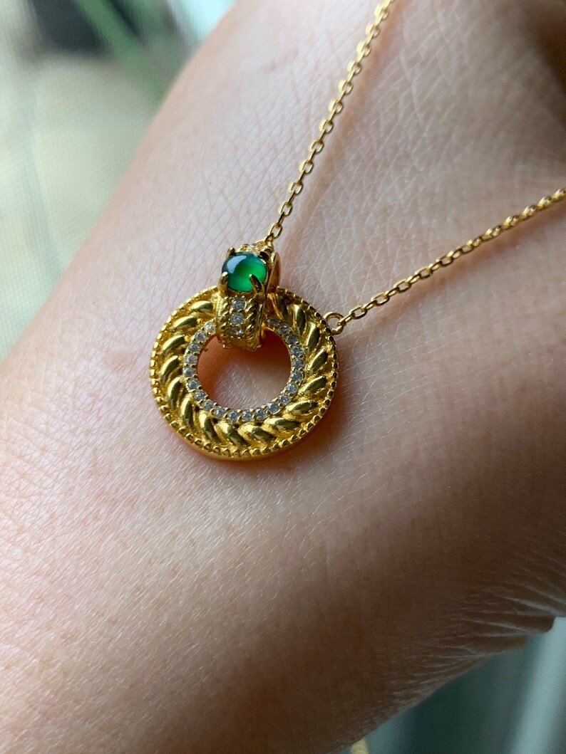 May include: A gold necklace with a circular pendant featuring a green gemstone and a row of clear stones around the edge.