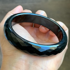 May include: A solid black bangle bracelet featuring a faceted exterior surface with multiple geometric cuts. The bracelet has a smooth, polished finish and a circular shape, showcasing a deep, dark color throughout the entire piece of jewelry.