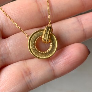 May include: A gold necklace with a circular pendant featuring a smaller, textured gold bar attached to the circle. The pendant has a detailed, textured edge.