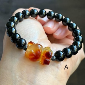 Könnte beinhalten: Ein schwarzes Perlenarmband mit einem zentralen orange-gelben Bärenanhänger. Das Armband wird in einer Hand gehalten, wobei die glänzenden, runden Perlen und der detaillierte Anhänger hervorgehoben werden.