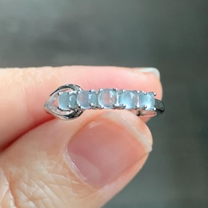 May include: A silver ring featuring five light blue oval gemstones. The gemstones are set in a row across the top of the ring. The ring is held in a hand, with the background out of focus.