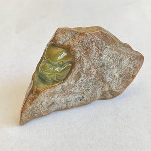 Puede incluir: Una piedra rugosa de forma triangular con un exterior texturizado, marrón y desgastado. Una sección lisa y pulida en la parte frontal revela un relieve tallado, translúcido y de color verde claro, de una criatura similar a un dragón.