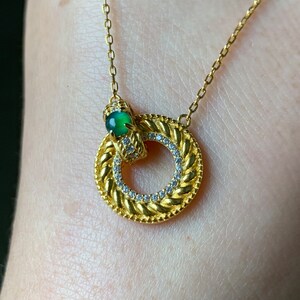 May include: A gold necklace with a circular pendant featuring a braided design and a small green gemstone. The pendant is accented with small clear stones.