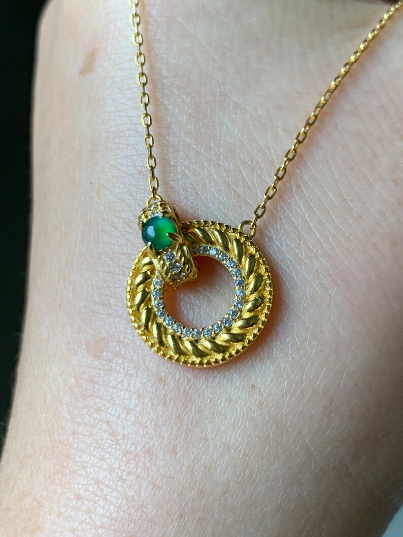 May include: A gold necklace with a circular pendant featuring a braided design and a small green gemstone. The pendant is accented with small clear stones.