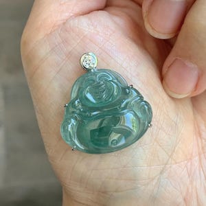 May include: A translucent, light green jade pendant in the shape of a laughing Buddha. The pendant is mounted on a silver setting and features a small, round diamond at the top. The jade has a smooth, polished surface.