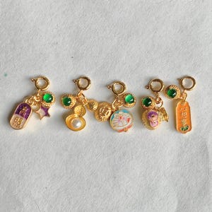 May include: Five gold-coloured charms with various designs, including a pill, a star, a pearl, a cartoon character, and a figure with wings. Each charm has a green gem and a small loop for attachment.