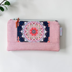 May include: A pink zippered pouch with a decorative mandala design. The pouch features a woven texture and a rectangular panel with a detailed, colorful mandala pattern. The zipper pull is orange. The words "cosie bag" are printed on the side.