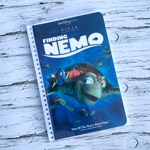 Peut inclure: Une jaquette de DVD du film Le Monde de Nemo avec un poisson bleu et orange nageant à côté d'une grosse tortue de mer verte sur un fond bleu. Le texte "Le Monde de Nemo" est en lettres blanches avec un contour bleu. Le texte "Un des meilleurs films de l'année." est en lettres blanches avec un contour noir.