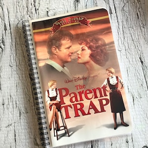 Puede incluir: Una libreta de espiral blanca con una portada que presenta el cartel de la película "La trampa de los padres" de la colección Walt Disney Vault. El cartel presenta a una pareja mirándose, una niña sentada en un taburete y una niña de pie con las manos en las caderas. El título de la película está en letras rojas grandes.