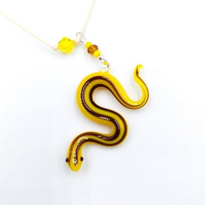 Könnte beinhalten: Ein gelber und brauner Glasschlangenanhänger an einer weißen Kordelkette. Die Schlange ist in einer 'S'-Form gekrümmt und hat eine glänzende Oberfläche.