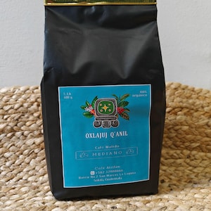 Könnte beinhalten: Schwarzer Kaffeepackung mit goldenem Deckel, beschriftet mit "OXLAJUJ Q'ANIL". Das türkisfarbene Etikett zeigt eine Maya-Grafik und den Text "Cafe Molido" und "MEDIANO". Die Packung ist mit 400 g und 100% Bio gekennzeichnet. Die Adresse des Cafés in Guatemala ist unten auf der Packung angegeben.