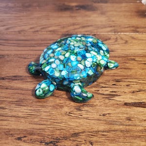 May include: A turtle-shaped decorative object featuring a shell and limbs filled with a mosaic of small, polished stones in various shades of blue, teal, green, and white, all encased in a smooth, clear, glossy resin material.