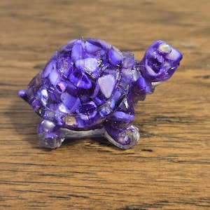 May include: A small, decorative turtle figurine. The turtle's shell is composed of small, purple and white pieces, giving it a mosaic appearance. The head, legs, and tail are a translucent purple. The figurine is resting on a wooden surface.