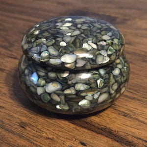 May include: A small, round decorative box with a lid. The box is filled with small, colorful stones in shades of green, white, and gray. The box is sitting on a wooden surface. The box is likely used for storing small items.
