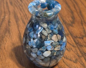 May include: A small, clear glass vase filled with blue and white stones. The vase has a rounded bottom and a slightly flared top.