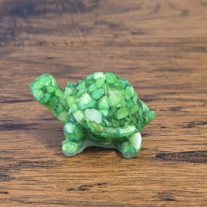 May include: A small, decorative turtle figurine. The turtle is primarily green with a mosaic-like shell pattern of small, rounded green and white pieces. The figurine has a smooth, glossy finish and is set against a wooden surface.